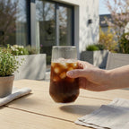 Glass med riller og cola. Ute på en terrasse holdt av en hånd. 