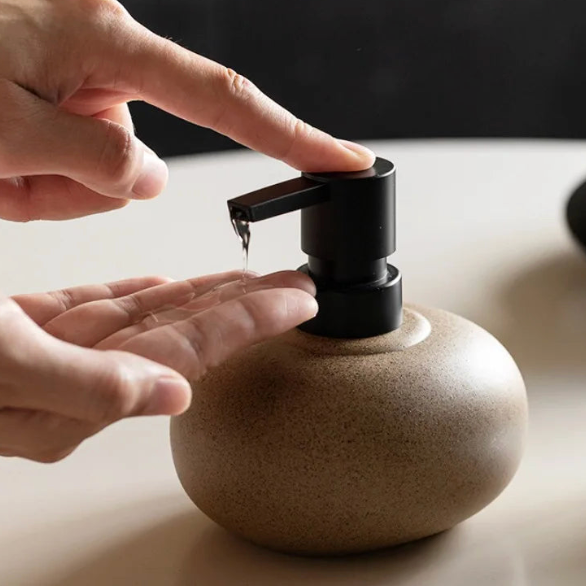 Lav, kuleformet såpedispenser i sandfarget keramikk med svart metallpumpe med en finger som presser ned.
